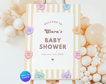 Könnte beinhalten: Ein weißes Willkommens-Schild mit gelben und weißen Streifen für eine Babyparty. Das Schild lautet "Willkommen zur Babyparty von Clara, 14. Februar 20XX". Das Schild ist mit bunten Bonbonherzen mit Sprüchen wie "Wahre Liebe", "Ich liebe dich", "Küss mich", "Sei mein", "Berausche mich", und "Du & Ich" verziert.