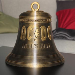 Könnte beinhalten: Eine bronzefarbene Metallglocke mit dem AC/DC-Logo und dem eingeprägten Schriftzug HELL'S BELL auf der Vorderseite. Die Glocke hat oben eine kreisförmige Aufhängung und am oberen Rand die Inschrift TAYLOR LOUGHBOROUGH ENGLAND.