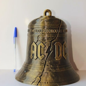 Peut inclure: Une cloche de couleur bronze avec le texte "AC/DC" et un éclair. La cloche présente une fissure sur le côté et le texte "LOUGHBOROUGH" est visible. Un stylo bleu est à côté de la cloche.