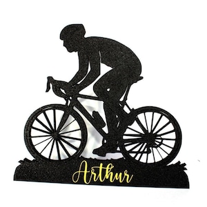 Può includere: Silhouette nera di un ciclista su una bici da corsa, con un casco. La bici ha ruote dettagliate e una borraccia. Il nome "Arthur" è scritto in oro sotto. L'immagine è su sfondo bianco.