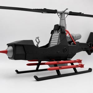 May include: A black toy helicopter featuring a silver nose cone with a red front-mounted cannon, a grey seat, and red missiles attached to the sides. The aircraft has black rotor blades, a silver rotor hub, and black landing skids on a plain background.