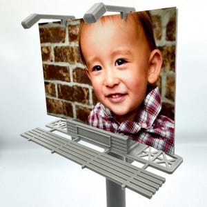 Könnte beinhalten: Ein graues Plastikmodell einer Werbetafel mit einem Foto eines lächelnden Kindes. Die Werbetafel hat zwei graue Lichter oben und einen grauen Metallrahmen.