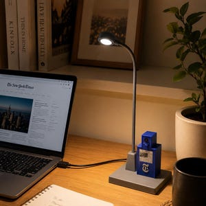 Può includere: Una lampada da scrivania grigia con collo flessibile e testa luminosa circolare, dotata di una base quadrata con un oggetto rettangolare blu attaccato. L'oggetto blu mostra una piccola grafica del giornale The New York Times e il logo T.