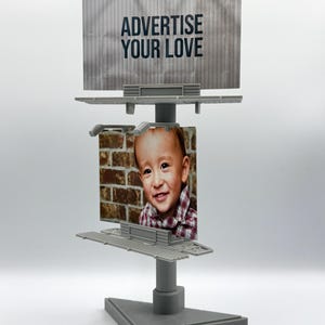 May include: A gray plastic miniature double-decker billboard display stand. The top sign features the text ADVERTISE YOUR LOVE in black letters on a gray background. The bottom sign displays a photograph of a smiling child in a plaid shirt.