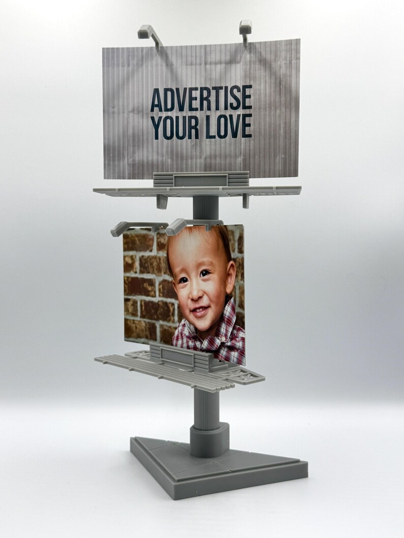 May include: A gray plastic miniature double-decker billboard display stand. The top sign features the text ADVERTISE YOUR LOVE in black letters on a gray background. The bottom sign displays a photograph of a smiling child in a plaid shirt.