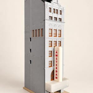 May include: A miniature model of a tall, narrow building with gray walls and orange-framed windows. A vertical white sign with the red text NEW AMSTERDAM is attached to the front facade above a translucent white marquee structure.