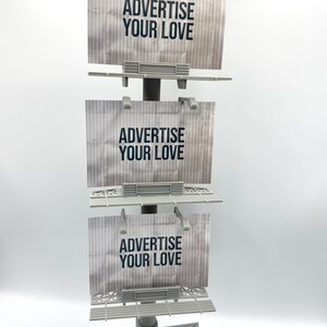 May include: A vertical display stand featuring three stacked rectangular billboard signs. Each sign has a light gray corrugated background with the text ADVERTISE YOUR LOVE printed in bold black letters. The structure includes small spotlights.