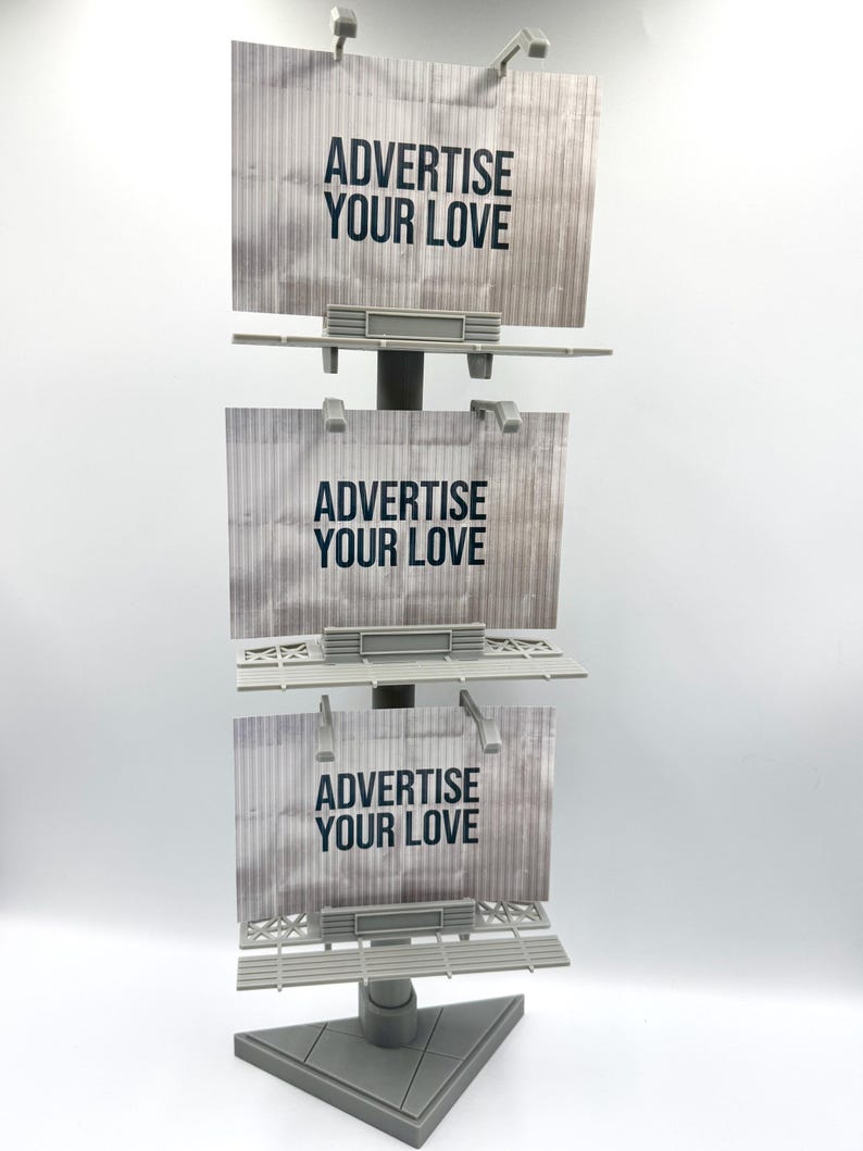 May include: A vertical display stand featuring three stacked rectangular billboard signs. Each sign has a light gray corrugated background with the text ADVERTISE YOUR LOVE printed in bold black letters. The structure includes small spotlights.