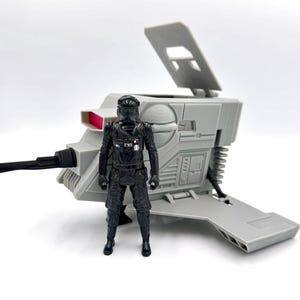 Könnte beinhalten: Eine schwarze Kunststoff-Actionfigur eines Piloten mit Helm und Fluganzug, die vor einem hellgrauen Kunststofffahrzeug mit offener Luke, einem roten rechteckigen Detail und einem schwarzen Kabel an der Seite des Aufbaus steht.