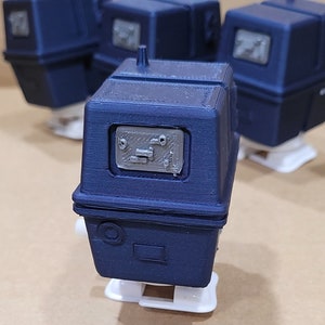 May include: Four dark blue 3D printed robot toys with white bases. The robots have a rectangular shape with a small dome on top. The front of the robots has a rectangular window with a grey design.