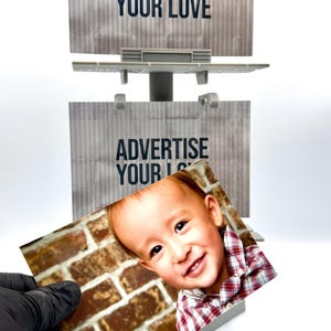 May include: A miniature gray plastic billboard structure featuring two stacked rectangular display panels with the text ADVERTISE YOUR LOVE printed in black. A rectangular photograph of a smiling child in a red plaid shirt is held in front.