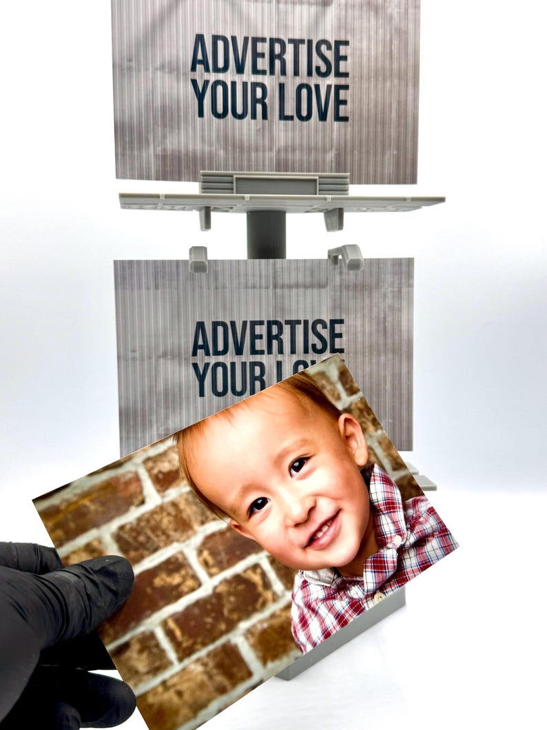 May include: A miniature gray plastic billboard structure featuring two stacked rectangular display panels with the text ADVERTISE YOUR LOVE printed in black. A rectangular photograph of a smiling child in a red plaid shirt is held in front.