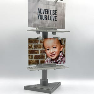 May include: A gray plastic double-sided billboard display stand featuring two rectangular panels. The top panel displays the text ADVERTISE YOUR LOVE in black letters, while the bottom panel holds a photograph of a smiling child.