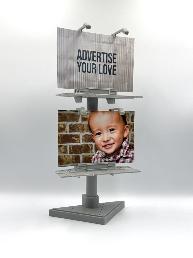 May include: A gray plastic double-sided billboard display stand featuring two rectangular panels. The top panel displays the text ADVERTISE YOUR LOVE in black letters, while the bottom panel holds a photograph of a smiling child.