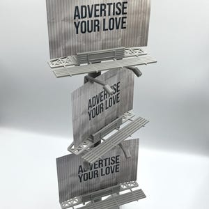 May include: A gray, three-tiered miniature billboard structure featuring a vertical pole and a triangular base. Each of the three rectangular panels displays the black text ADVERTISE YOUR LOVE against a textured, light gray background.