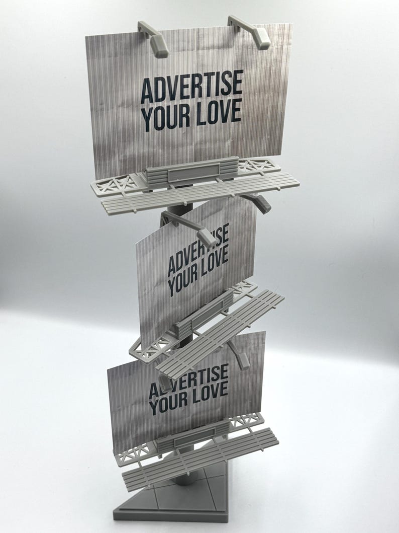 May include: A gray, three-tiered miniature billboard structure featuring a vertical pole and a triangular base. Each of the three rectangular panels displays the black text ADVERTISE YOUR LOVE against a textured, light gray background.