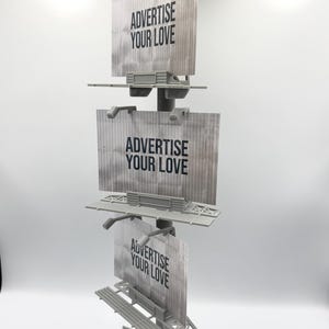 May include: A gray vertical display stand featuring three stacked rectangular billboard signs. Each sign has a textured surface and displays the black text ADVERTISE YOUR LOVE. The structure is supported by a central pole and a triangular base.