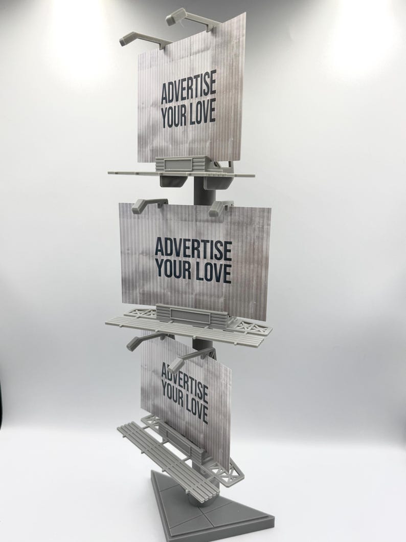 May include: A gray vertical display stand featuring three stacked rectangular billboard signs. Each sign has a textured surface and displays the black text ADVERTISE YOUR LOVE. The structure is supported by a central pole and a triangular base.