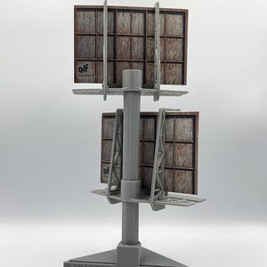 May include: A gray plastic model of a double-sided billboard structure with a central pole and triangular base. The base features the text "GreeblerElf 2025". Each billboard face displays a grid pattern with a rusted metal texture and the logo "elf".
