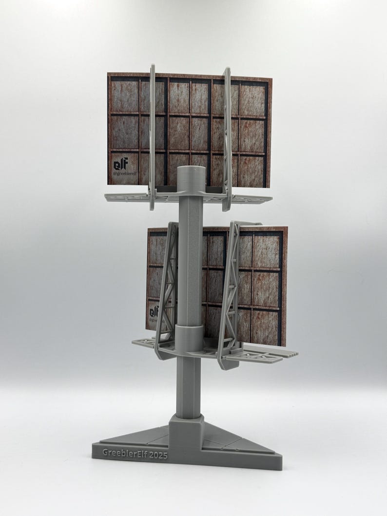 May include: A gray plastic model of a double-sided billboard structure with a central pole and triangular base. The base features the text "GreeblerElf 2025". Each billboard face displays a grid pattern with a rusted metal texture and the logo "elf".