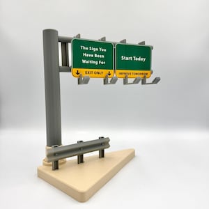 May include: A miniature highway sign with two green panels. One panel reads "The Sign You Have Been Waiting For" and the other says "Start Today." The base is tan, with a gray guardrail and support post. The sign is a novelty item.