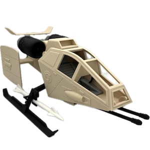 May include: Tan toy helicopter with black rotors, skis, and a clear cockpit. Two white missiles are attached to the black frame. The toy has a detailed design, including panel lines and a small window on top.