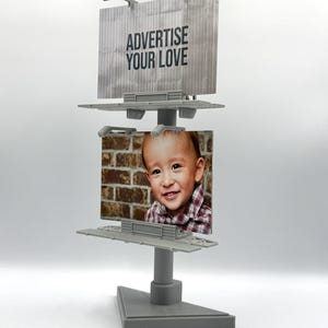 May include: A gray miniature billboard with the text "ADVERTISE YOUR LOVE" on top. The bottom section displays a photo of a smiling child in a plaid shirt. The billboard is on a gray stand.