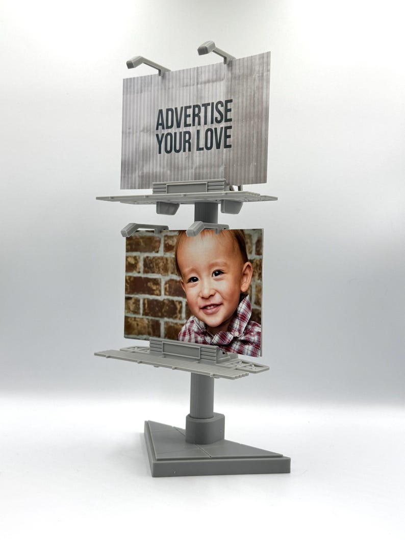May include: A gray plastic miniature double-sided billboard display stand featuring two rectangular panels. The top panel displays the text ADVERTISE YOUR LOVE on a gray background, while the bottom panel features a photograph of a smiling child.