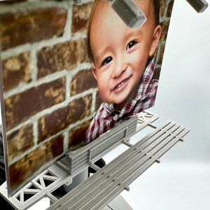 May include: A miniature gray plastic billboard structure featuring a printed photograph of a smiling child wearing a red and white plaid shirt against a brick wall background. The display is mounted on a gray base with a tiered platform design.