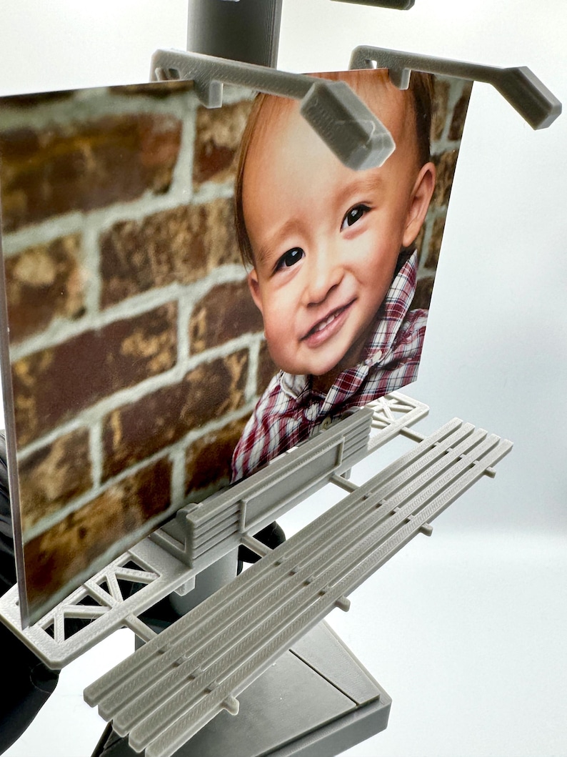 May include: A miniature gray plastic billboard structure featuring a printed photograph of a smiling child wearing a red and white plaid shirt against a brick wall background. The display is mounted on a gray base with a tiered platform design.