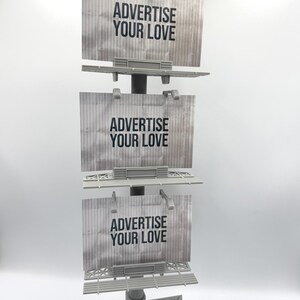 May include: A gray vertical display stand featuring three stacked rectangular billboard signs. Each sign has a textured metallic appearance and displays the black text ADVERTISE YOUR LOVE. The structure is supported by a triangular base.