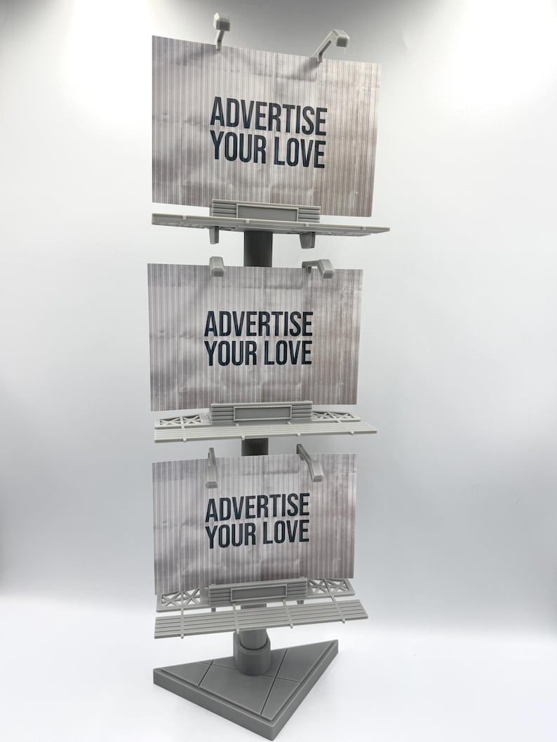 May include: A gray vertical display stand featuring three stacked rectangular billboard signs. Each sign has a textured metallic appearance and displays the black text ADVERTISE YOUR LOVE. The structure is supported by a triangular base.