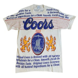 Puede incluir: Una camiseta blanca con un patrón de rayas azules y rojas. La camiseta presenta un gran logotipo de Coors Beer con una escena de montaña y dos leones amarillos. El texto "Fresh from... is Brewed with All Natural Ingredients for a Clean, Smooth, Fresh from the Rockies Taste." está impreso en la camiseta.