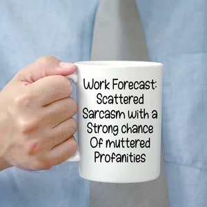 May include: White ceramic mug with the text "Work Forecast: Scattered Sarcasm with a Strong chance Of muttered Profanities." The mug is held by a person wearing a blue shirt and a gray tie. The mug is a standard size.