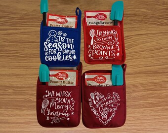 Op de afbeelding: Vier rode en blauwe ovenwantjes met zakken om bakbenodigdheden in te bewaren. Elk wantje heeft een ander kerstthema en tekst. De wantjes zijn versierd met witte tekst en afbeeldingen. De tekst op de wantjes is: "Tis the season for baking cookies", "Trying to earn brownie points", "We whisk you a Merry Christmas", en "Salt, sugar, milk, vanilla, cocoa, butter, flour, cinnamon".