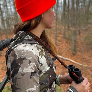 May include: A person wearing a camouflage jacket and an orange beanie is holding binoculars with a green and orange strap. The person is sitting in a tree stand and looking out into the woods.