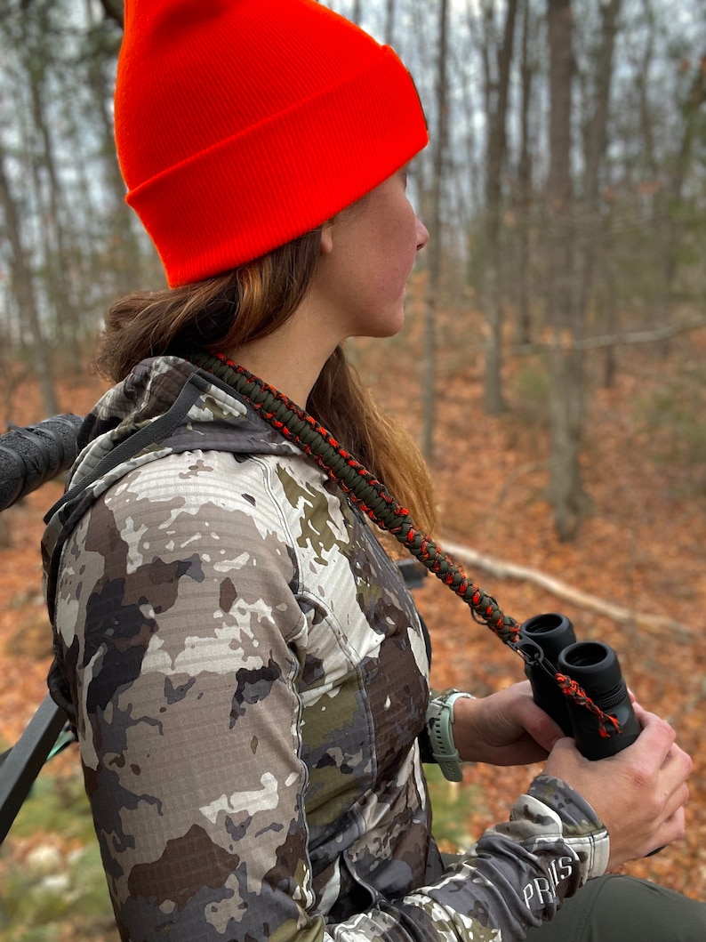 May include: A person wearing a camouflage jacket and an orange beanie is holding binoculars with a green and orange strap. The person is sitting in a tree stand and looking out into the woods.
