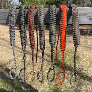 May include: Eight different paracord slings in various camouflage patterns, including orange and brown, hanging on a wooden fence.