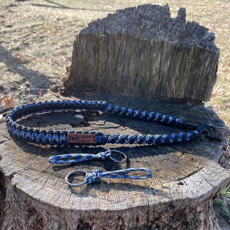 May include: A black and blue paracord leash with a brown leather tag that reads "The Upland Creative". The leash is on a wooden surface with two matching paracord keychains.