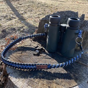 May include: A black and blue paracord strap with a brown leather tag that reads "The Upland Creative". The strap is wrapped around a pair of black binoculars.