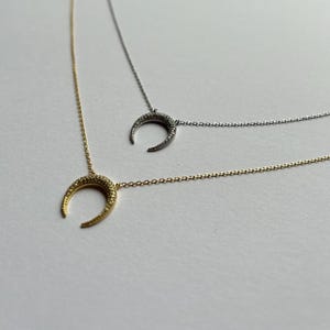 May include: Two delicate necklaces with crescent moon pendants. The top necklace is silver with a silver crescent moon pendant, and the bottom necklace is gold with a gold crescent moon pendant. Both pendants are encrusted with small, sparkling stones.
