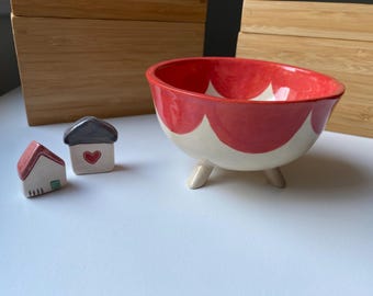 May include: A ceramic bowl with a red and cream scalloped design, supported by three small legs. Two miniature ceramic houses with red and gray roofs and heart details are also present. The items are on a white surface.