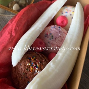 May include: A set of three ice cream-themed candles in a red and gold gift box. The candles are shaped like ice cream cones and are decorated with sprinkles and cherries. The box has the website www.kluxewaxstudio.com printed on it.