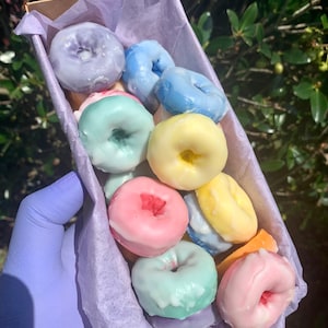 May include: A box filled with colorful, miniature donuts. The donuts are glazed in pastel colors like blue, pink, yellow, and green. The box is lined with purple tissue paper, and held by a gloved hand.