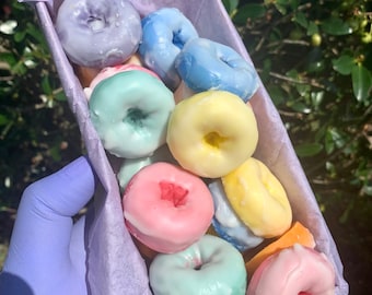 May include: A box filled with colorful, miniature donuts. The donuts are glazed in pastel colors like blue, pink, yellow, and green. The box is lined with purple tissue paper, and held by a gloved hand.