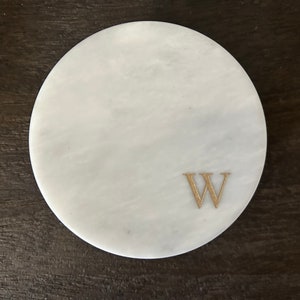 May include: A round, white marble coaster with a gold-colored letter "W" etched on the surface. The coaster has a smooth, polished finish and is designed to protect surfaces from drink condensation.