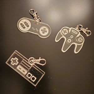 May include: Three clear acrylic keychains shaped like video game controllers. The keychains are attached to silver metal rings. The controllers are a Nintendo Entertainment System controller, a Super Nintendo Entertainment System controller, and a Nintendo 64 controller.