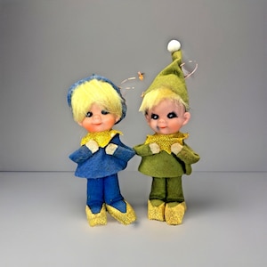 May include: Two vintage elf dolls. One doll is wearing a blue outfit with a yellow collar and shoes, and a blue hat. The other doll is wearing a green outfit with a yellow collar and shoes, and a green hat. Both dolls have yellow hair.
