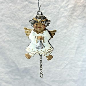 May include: A decorative angel ornament with a white dress, gold wings, and a silver bell. The angel has dark skin, a smiling face, and a gold halo. A silver chain hangs from the bottom of the ornament.