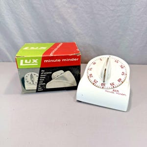 May include: A white Lux Minute Minder kitchen timer with a central dial and red numerical markings, accompanied by its original cardboard box featuring green, red, and black panels with the text Lux minute minder and all purpose timer.